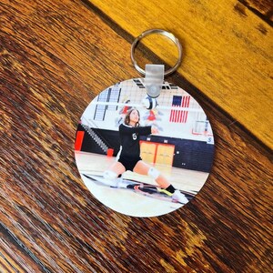 May include: A round keychain with a photo of a volleyball player in a black and white uniform, jumping to spike the ball. The player is wearing a number 5 on their jersey. The photo is taken in a gymnasium with a volleyball net in the background.