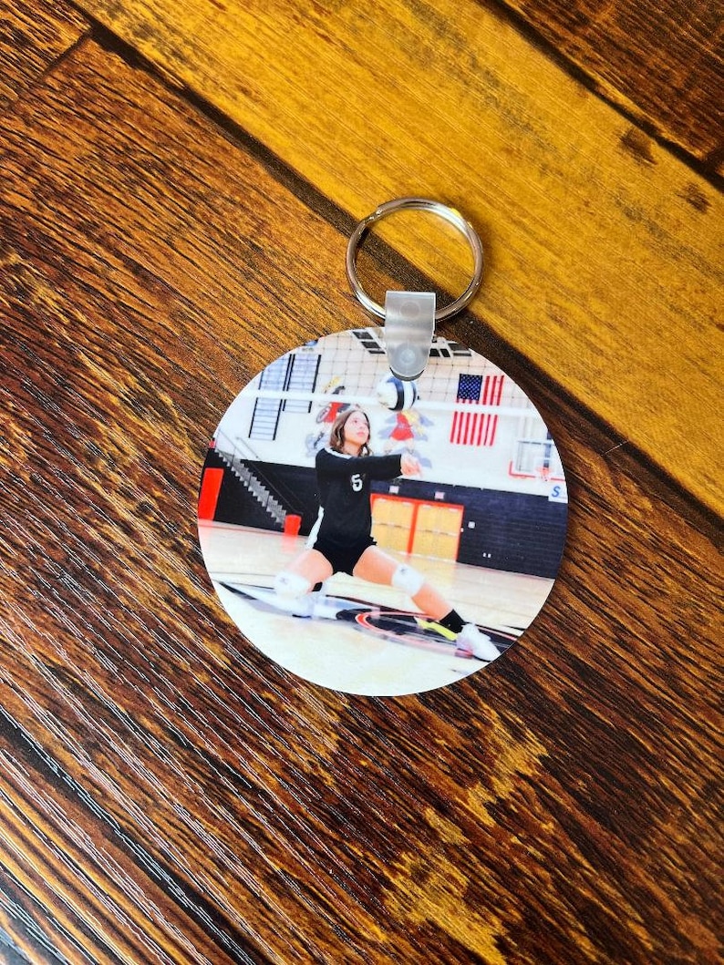 May include: A round keychain with a photo of a volleyball player in a black and white uniform, jumping to spike the ball. The player is wearing a number 5 on their jersey. The photo is taken in a gymnasium with a volleyball net in the background.