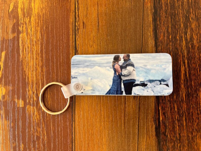 May include: A keychain with a photo of a couple standing on a snowy beach. The couple is wearing winter clothing and is looking at each other. The photo is printed on a white background.