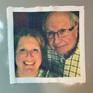 May include: A close-up photo of a couple, a man and a woman, smiling at the camera. The man is wearing a plaid shirt and glasses. The woman is wearing a patterned shirt.