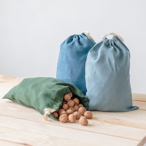 Könnte beinhalten: Zwei blaue wiederverwendbare Stoffbeutel und ein grüner wiederverwendbarer Stoffbeutel gefüllt mit Walnüssen auf einem hellen Holztisch.