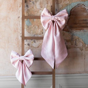 May include: Two pink satin bows tied with a loop on a wooden ladder leaning against a textured wall.