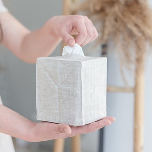 May include: A square tissue box cover made of light beige, textured fabric. The cover features a clean, structured design with visible seams along the edges and a central opening at the top from which a single white tissue is being pulled.