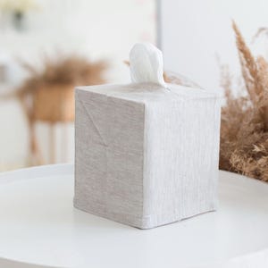 May include: A square tissue box cover made of light beige, textured fabric. The cover features a simple, clean design with a central opening at the top, through which a single white tissue is pulled upward, extending slightly above the box surface.