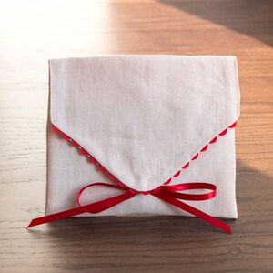 May include: A white fabric envelope with a red scalloped edge and a red ribbon tied in a bow.