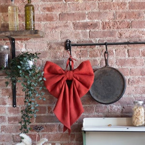 May include: A large, red fabric bow hangs from a hook on a brick wall. The bow is made of a linen-like material and has a rustic, farmhouse style.