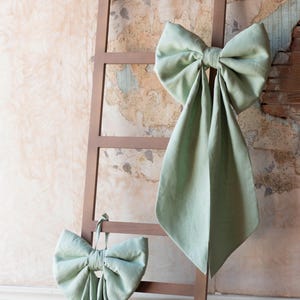 May include: Two large, sage green fabric bows are displayed on a brown wooden ladder. One bow is tied to the ladder's rungs, while the other is draped over the top. The background features a distressed wall.