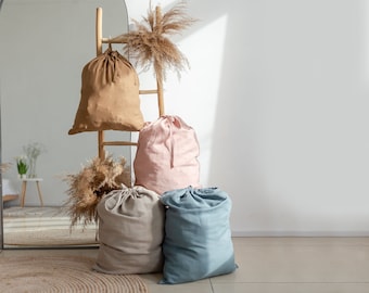 May include: Four fabric storage bags in different colours: brown, pink, beige, and blue. The bags are on a wooden ladder with a mirror behind them. The bags are made of a soft, natural fabric and have a drawstring closure.