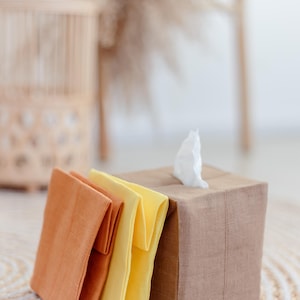 K&ouml;nnte beinhalten: Ein quadratischer Taschentuchbox-Bezug aus braunem Stoff mit einem einzelnen wei&szlig;en Taschentuch, das oben herausragt, begleitet von zwei gefalteten Stoffservietten in gebranntem Orange und Hellgelb, alle mit sichtbarer Webstruktur.