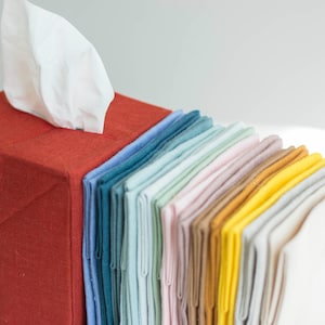 K&ouml;nnte beinhalten: Ein quadratischer, rostfarbener Stoff&uuml;berzug f&uuml;r eine Taschentuchbox, aus dem oben ein wei&szlig;es Taschentuch herausragt. Neben der Box befindet sich ein vertikaler Stapel gefalteter Stoffservietten in einem Farbverlauf aus Blau, Petrol, Mint, Rosa, Beige, Gelb und Wei&szlig;.