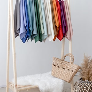 May include: A wooden clothing rack with a variety of colored linen cloths hanging on it. The rack has a shelf and a white faux fur rug on the shelf. A woven basket bag is sitting on the rug.