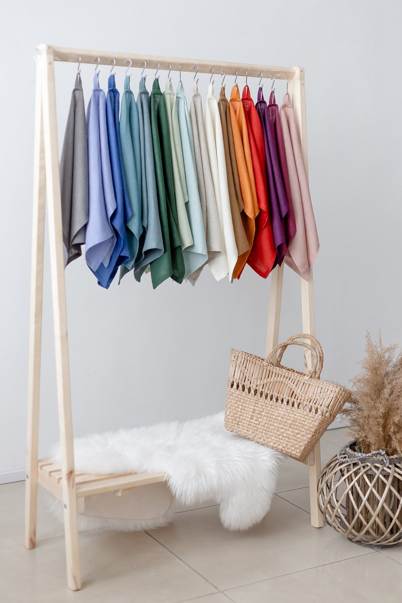 May include: A wooden clothing rack with a variety of colored linen cloths hanging on it. The rack has a shelf and a white faux fur rug on the shelf. A woven basket bag is sitting on the rug.