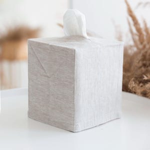 May include: A square tissue box cover made of light beige, textured fabric. The cover features a top opening with a single white tissue protruding from the center. The fabric has a visible weave pattern and clean, straight stitched edges.
