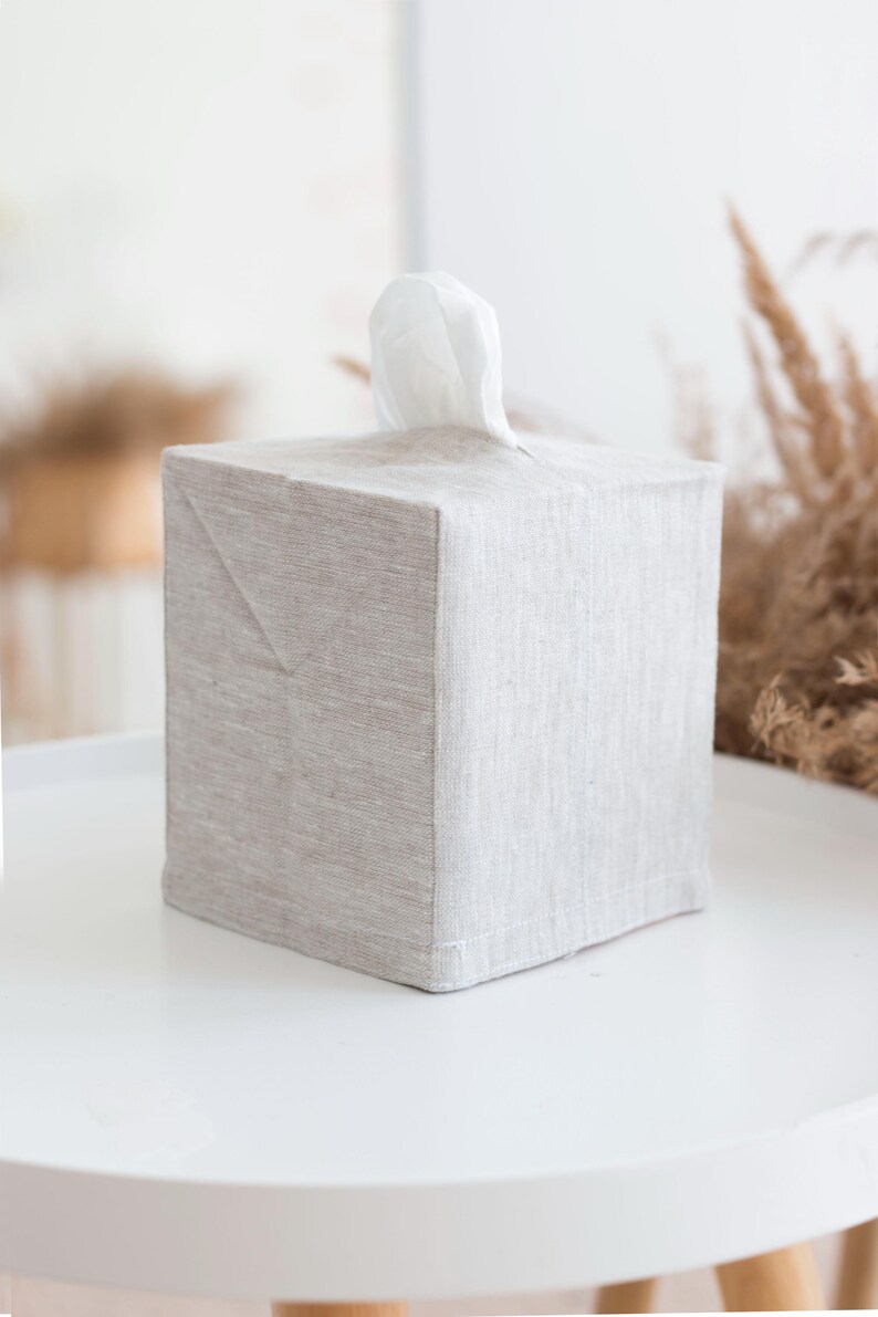 May include: A square tissue box cover made of light beige, textured fabric. The cover features a top opening with a single white tissue protruding from the center. The fabric has a visible weave pattern and clean, straight stitched edges.