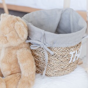 May include: A grey linen-lined wicker basket with a white fabric handle. The basket is sitting on a white fluffy surface next to a brown teddy bear.