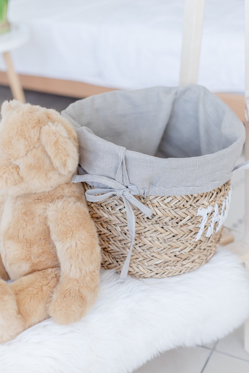 May include: A grey linen-lined wicker basket with a white fabric handle. The basket is sitting on a white fluffy surface next to a brown teddy bear.
