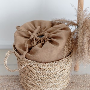 May include: A brown linen drawstring bag sits inside a woven wicker basket with a handle. The basket is on a woven rug.