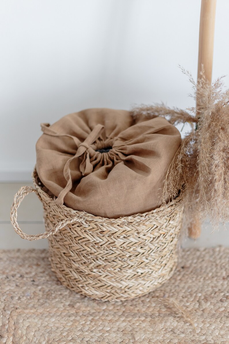 May include: A brown linen drawstring bag sits inside a woven wicker basket with a handle. The basket is on a woven rug.