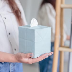 K&ouml;nnte beinhalten: Ein quadratischer, hellblauer Taschentuchbox-&Uuml;berzug aus Stoff mit sichtbarer Webstruktur. Der &Uuml;berzug verf&uuml;gt &uuml;ber eine &Ouml;ffnung an der Oberseite, aus der ein einzelnes wei&szlig;es Taschentuch in der Mitte herausragt. Die Form ist w&uuml;rfelf&ouml;rmig.