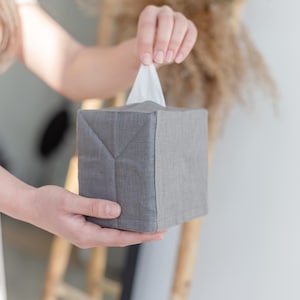 K&ouml;nnte beinhalten: Ein quadratischer Taschentuchbox-&Uuml;berzug aus grauem Stoff mit sichtbarer Webstruktur. Der &Uuml;berzug verf&uuml;gt &uuml;ber eine obere &Ouml;ffnung, aus der ein einzelnes wei&szlig;es Taschentuch in der Mitte herausragt. Der Stoff ist an den Ecken gefaltet.