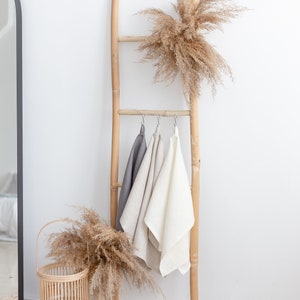 May include: A wooden ladder with three linen towels hanging from it. The ladder is decorated with dried pampas grass. A woven basket with dried pampas grass sits on a woven rug.