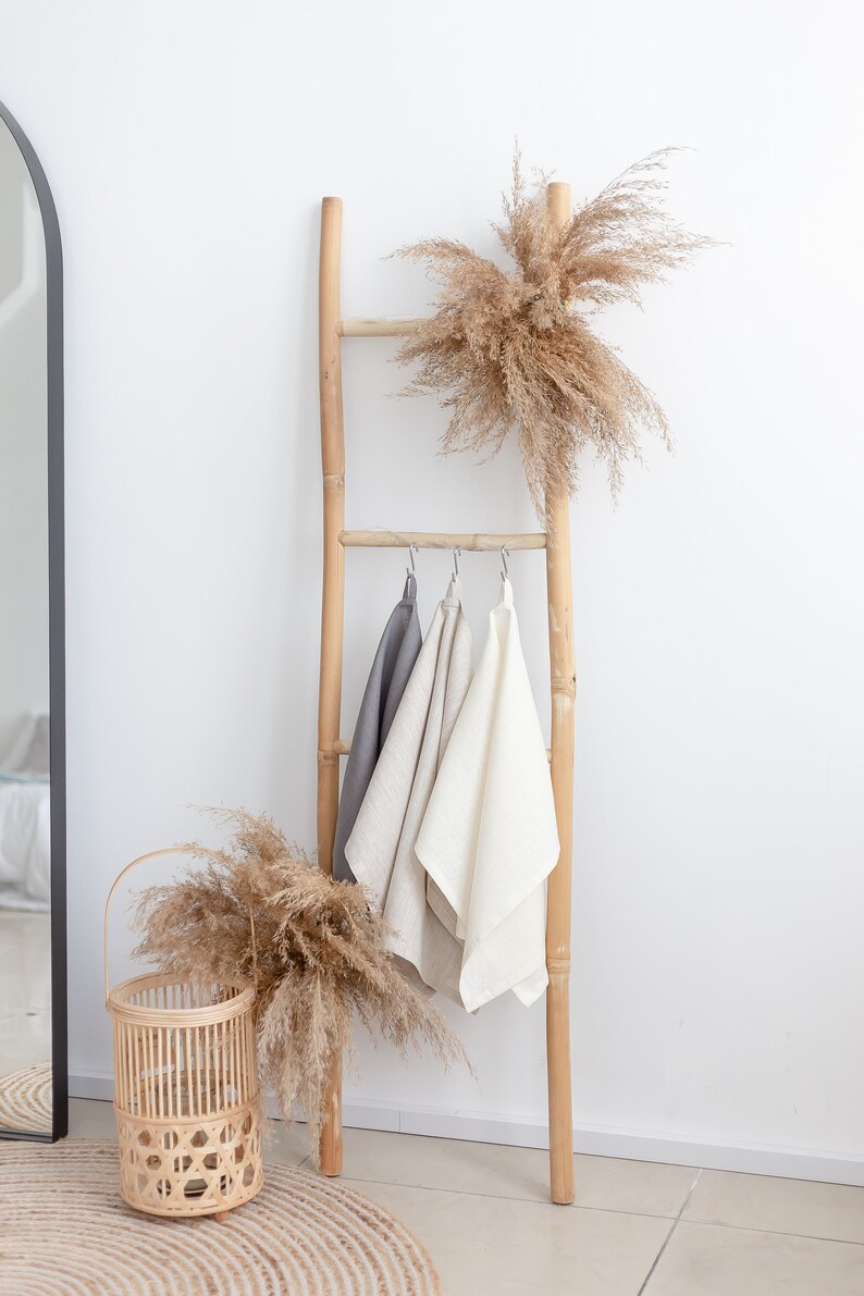 May include: A wooden ladder with three linen towels hanging from it. The ladder is decorated with dried pampas grass. A woven basket with dried pampas grass sits on a woven rug.