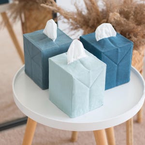 May include: Three square tissue box covers made of fabric in varying shades of blue, including light blue, medium blue, and dark blue. Each cover features a top opening with a white tissue protruding from the center and visible folded seams.
