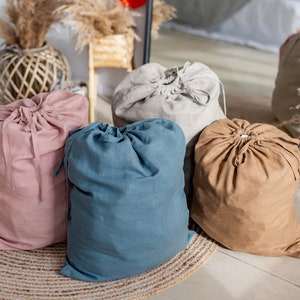 May include: Four drawstring linen bags in different colours: pink, blue, beige, and grey. The bags are sitting on a woven rug.