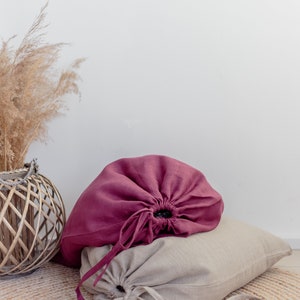 May include: Two linen drawstring bags, one burgundy and one beige, are sitting on a woven rug. The bags are gathered at the top and tied with matching cords.