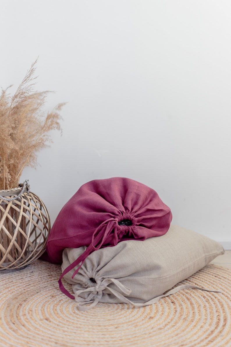 May include: Two linen drawstring bags, one burgundy and one beige, are sitting on a woven rug. The bags are gathered at the top and tied with matching cords.