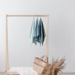 May include: A wooden clothing rack with three light blue linen towels hanging on it. A white faux fur rug is on the bottom shelf. A wicker basket and a dried pampas grass arrangement are on the floor.