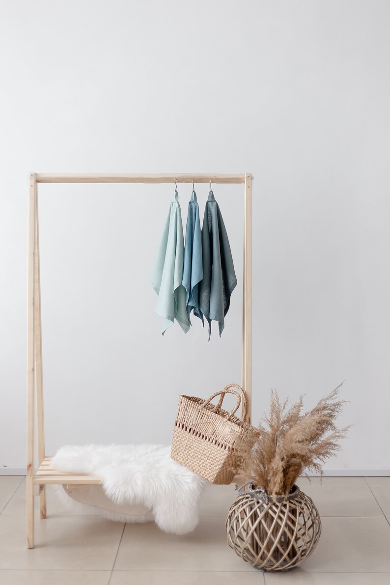 May include: A wooden clothing rack with three light blue linen towels hanging on it. A white faux fur rug is on the bottom shelf. A wicker basket and a dried pampas grass arrangement are on the floor.
