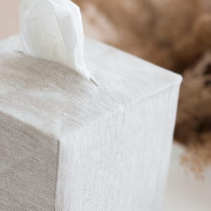 May include: A square tissue box cover made of light beige, textured fabric. The cover features a central rectangular opening at the top, through which a single white tissue is pulled upward. The fabric has a visible weave pattern throughout.