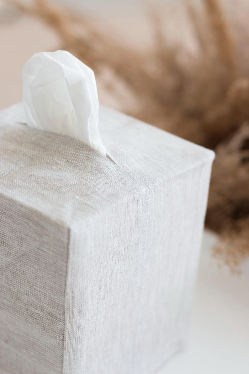 May include: A square tissue box cover made of light beige, textured fabric. The cover features a central rectangular opening at the top, through which a single white tissue is pulled upward. The fabric has a visible weave pattern throughout.