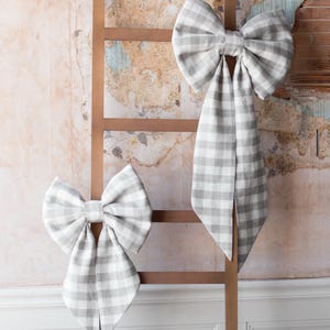 May include: Two large, gray and white gingham fabric bows are displayed on a wooden ladder against a textured wall. The bows have long tails and are tied in the center. The background has a vintage, distressed look.