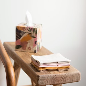 K&ouml;nnte beinhalten: Ein quadratischer Taschentuchbox-Bezug mit einem bunten abstrakten Muster aus rosa, senfgelben, gr&uuml;nen und schwarzen Formen, kombiniert mit einem Stapel gefalteter Stoffservietten in passenden Farbt&ouml;nen wie Creme, Hellrosa, Senf und Braun.