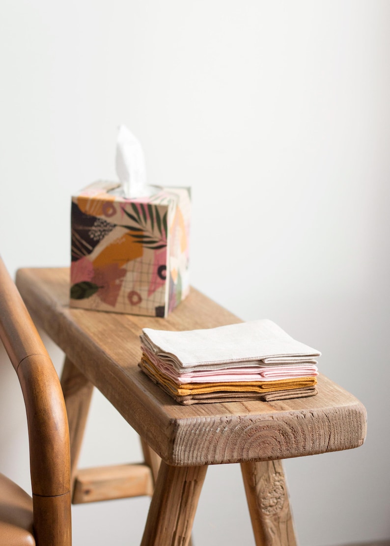 K&ouml;nnte beinhalten: Ein quadratischer Taschentuchbox-Bezug mit einem bunten abstrakten Muster aus rosa, senfgelben, gr&uuml;nen und schwarzen Formen, kombiniert mit einem Stapel gefalteter Stoffservietten in passenden Farbt&ouml;nen wie Creme, Hellrosa, Senf und Braun.