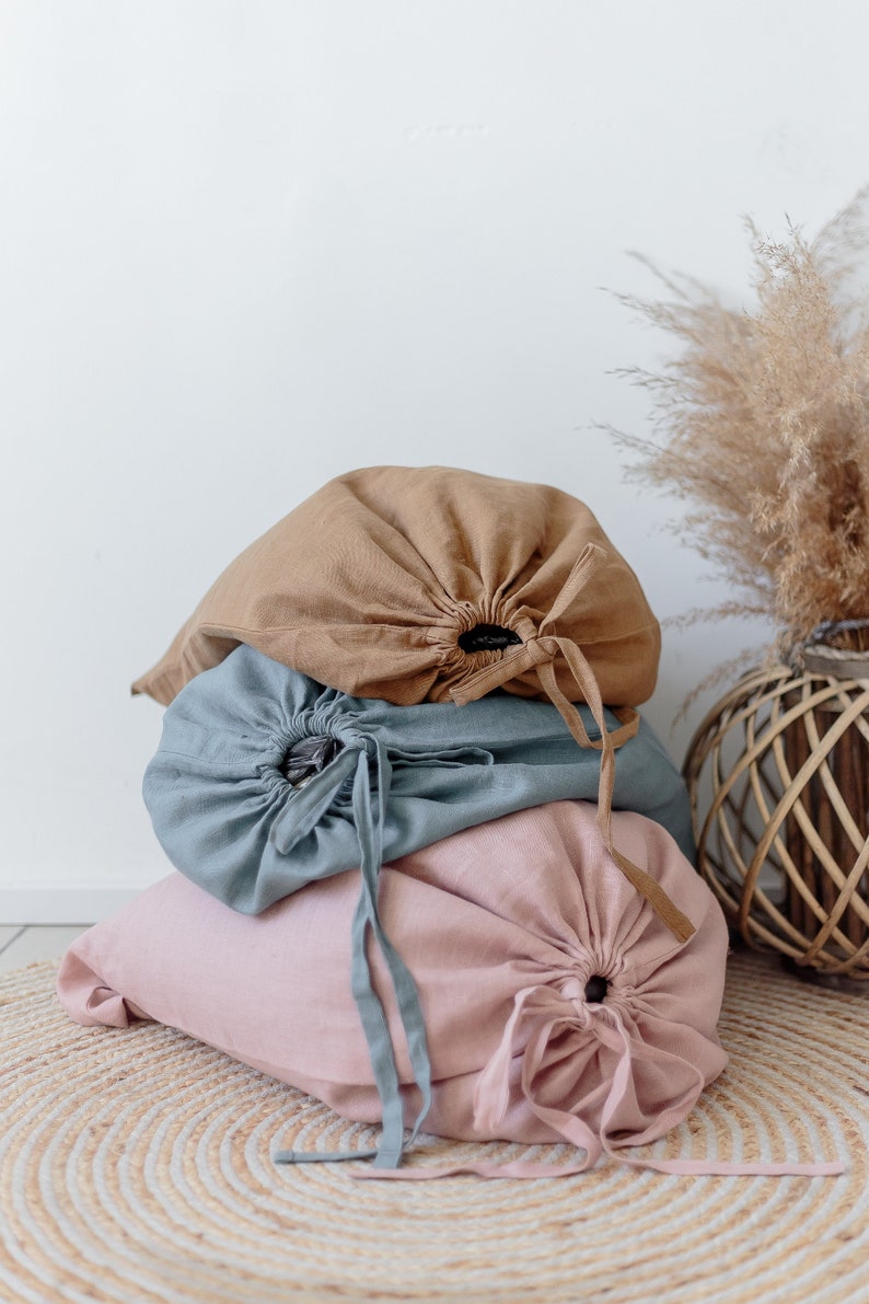 May include: Three fabric storage bags in different colours: brown, blue, and pink. The bags are made of linen and have a drawstring closure. The bags are stacked on top of each other and are sitting on a woven rug.