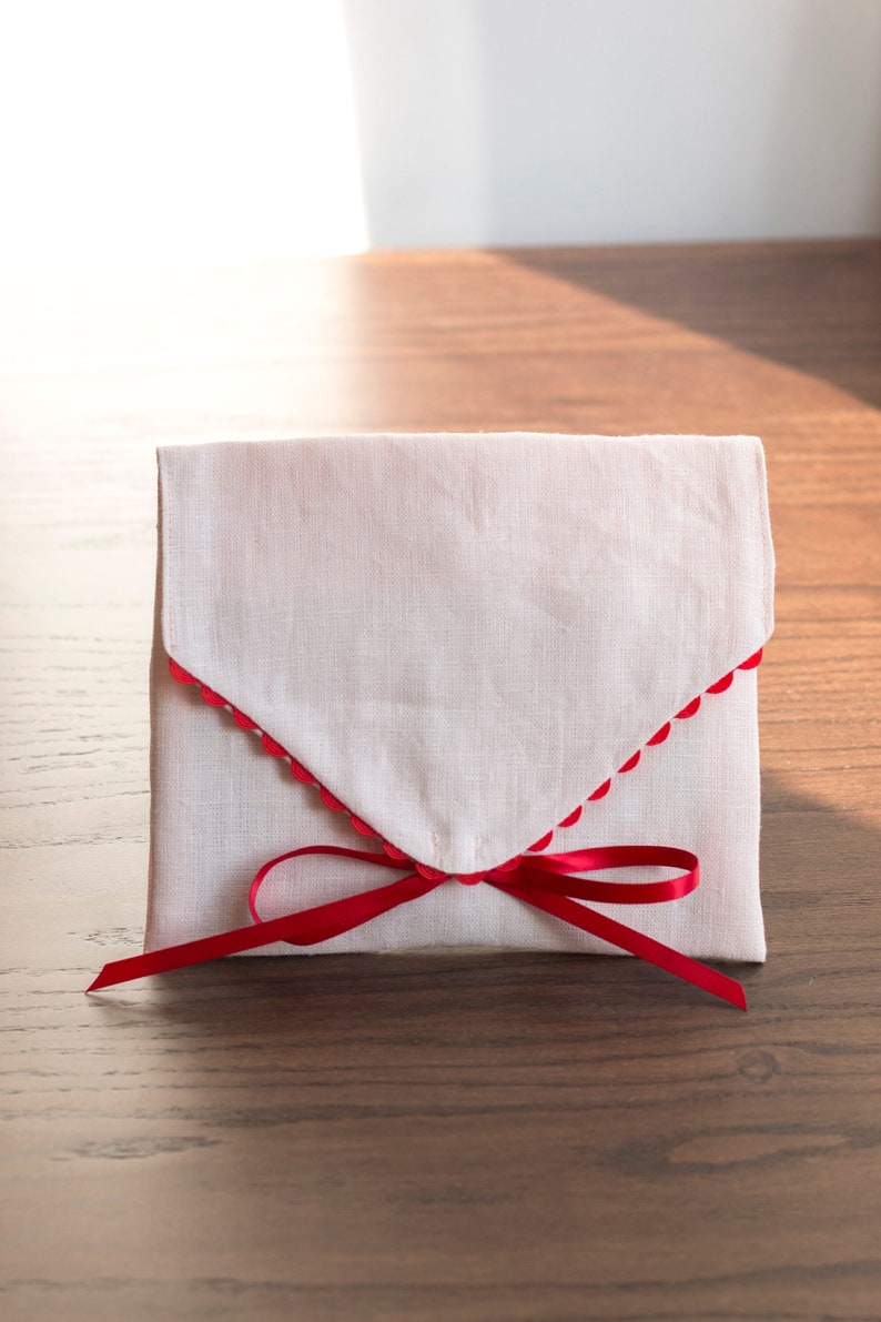May include: A white fabric envelope with a red scalloped edge and a red ribbon tied in a bow.