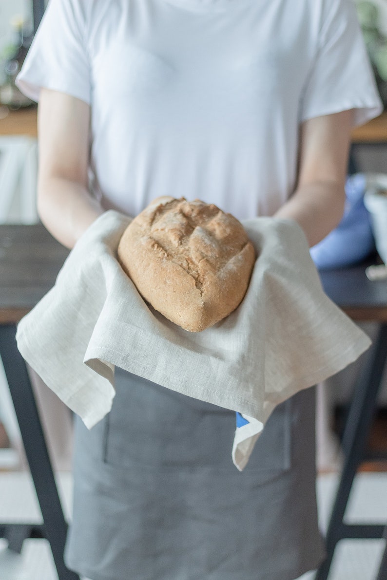 May include: A person holding a loaf of freshly baked bread on a white linen tea towel.