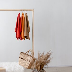 May include: A wooden clothing rack with three scarves hanging on it. The scarves are red, orange, and brown. There is a wicker basket and a dried grass bouquet in the foreground.