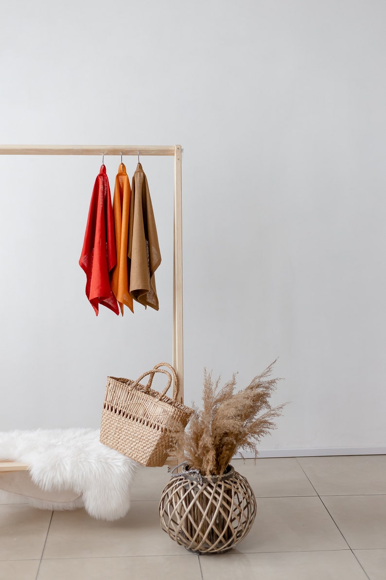 May include: A wooden clothing rack with three scarves hanging on it. The scarves are red, orange, and brown. There is a wicker basket and a dried grass bouquet in the foreground.