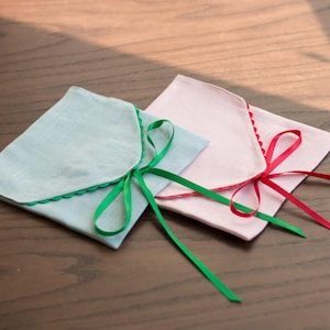 May include: Two fabric gift bags with scalloped edges. One bag is light blue with a green ribbon, and the other is pink with a red ribbon.