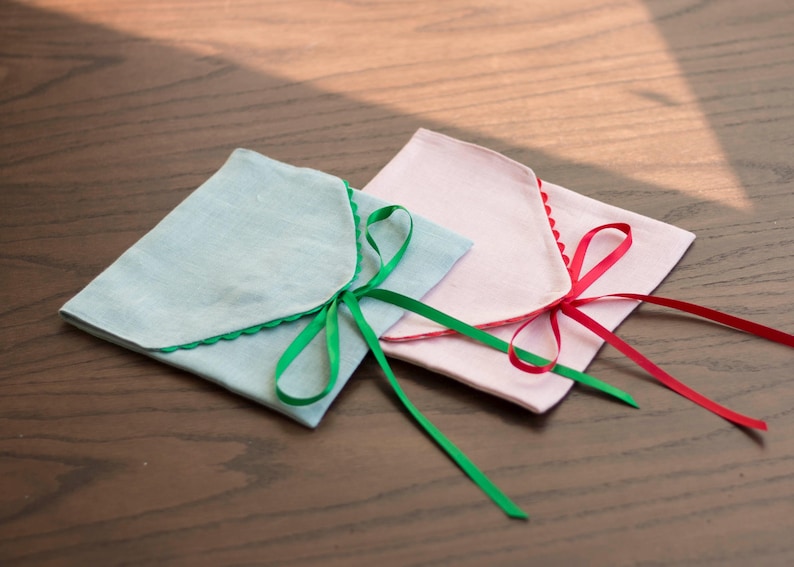May include: Two fabric gift bags with scalloped edges. One bag is light blue with a green ribbon, and the other is pink with a red ribbon.