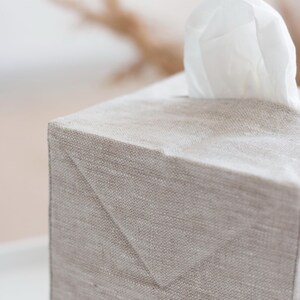 May include: A rectangular tissue box cover made of light beige, textured fabric. The cover features a clean, structured design with visible seams and a triangular fold detail at the top corner. A single white tissue protrudes from the top opening.