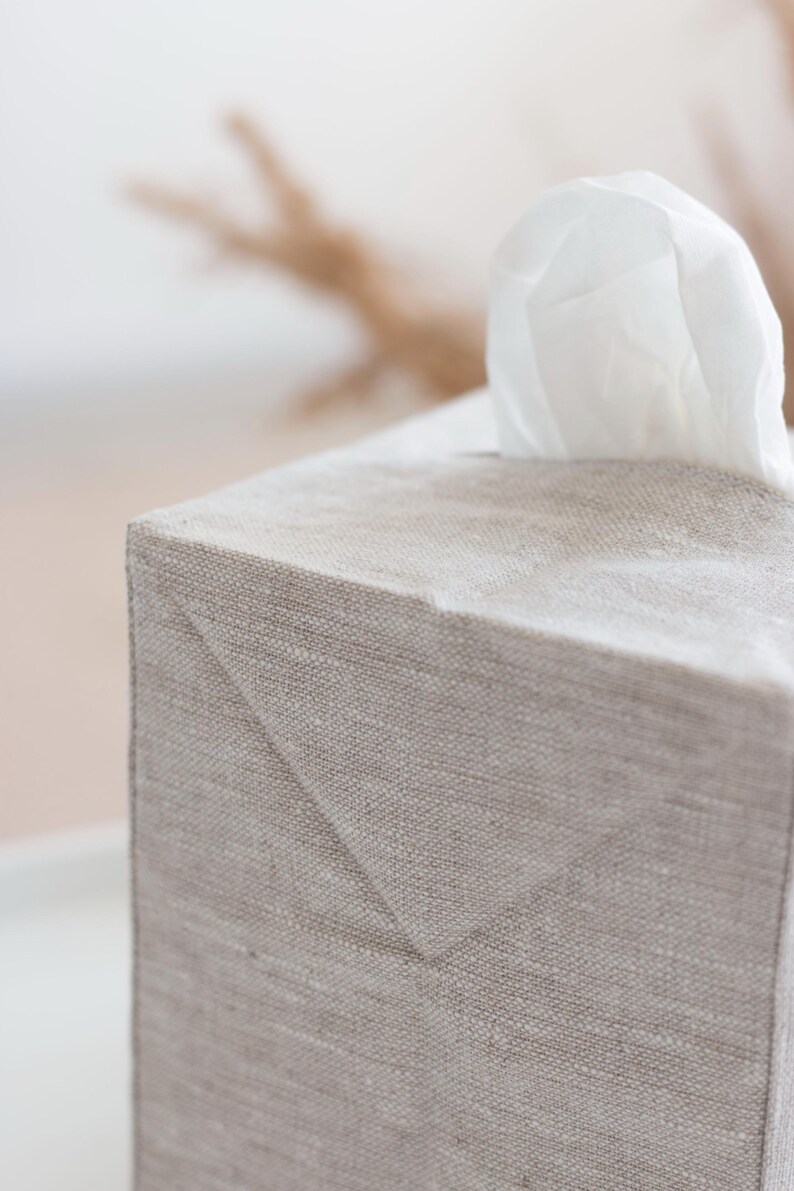 May include: A rectangular tissue box cover made of light beige, textured fabric. The cover features a clean, structured design with visible seams and a triangular fold detail at the top corner. A single white tissue protrudes from the top opening.