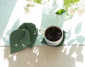 May include: A white ceramic mug filled with dark coffee sits on a green leaf-shaped coaster. Two additional green leaf-shaped coasters are to the left. The scene is set on a light blue surface with sunlight and floral accents.