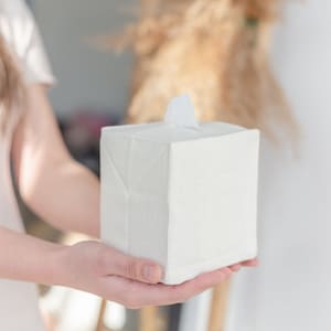 K&ouml;nnte beinhalten: Ein quadratischer, cremewei&szlig;er Gewebe-Taschentuchbox-&Uuml;berzug mit einer strukturierten, leinenartigen Optik. Der &Uuml;berzug zeichnet sich durch ein schlichtes Design mit gefalteten Ecken und einer mittigen, rechteckigen &Ouml;ffnung an der Oberseite aus.