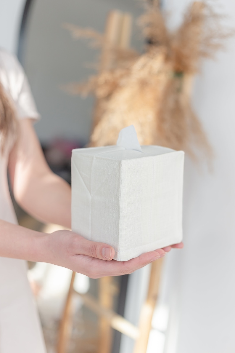 K&ouml;nnte beinhalten: Ein quadratischer, cremewei&szlig;er Gewebe-Taschentuchbox-&Uuml;berzug mit einer strukturierten, leinenartigen Optik. Der &Uuml;berzug zeichnet sich durch ein schlichtes Design mit gefalteten Ecken und einer mittigen, rechteckigen &Ouml;ffnung an der Oberseite aus.