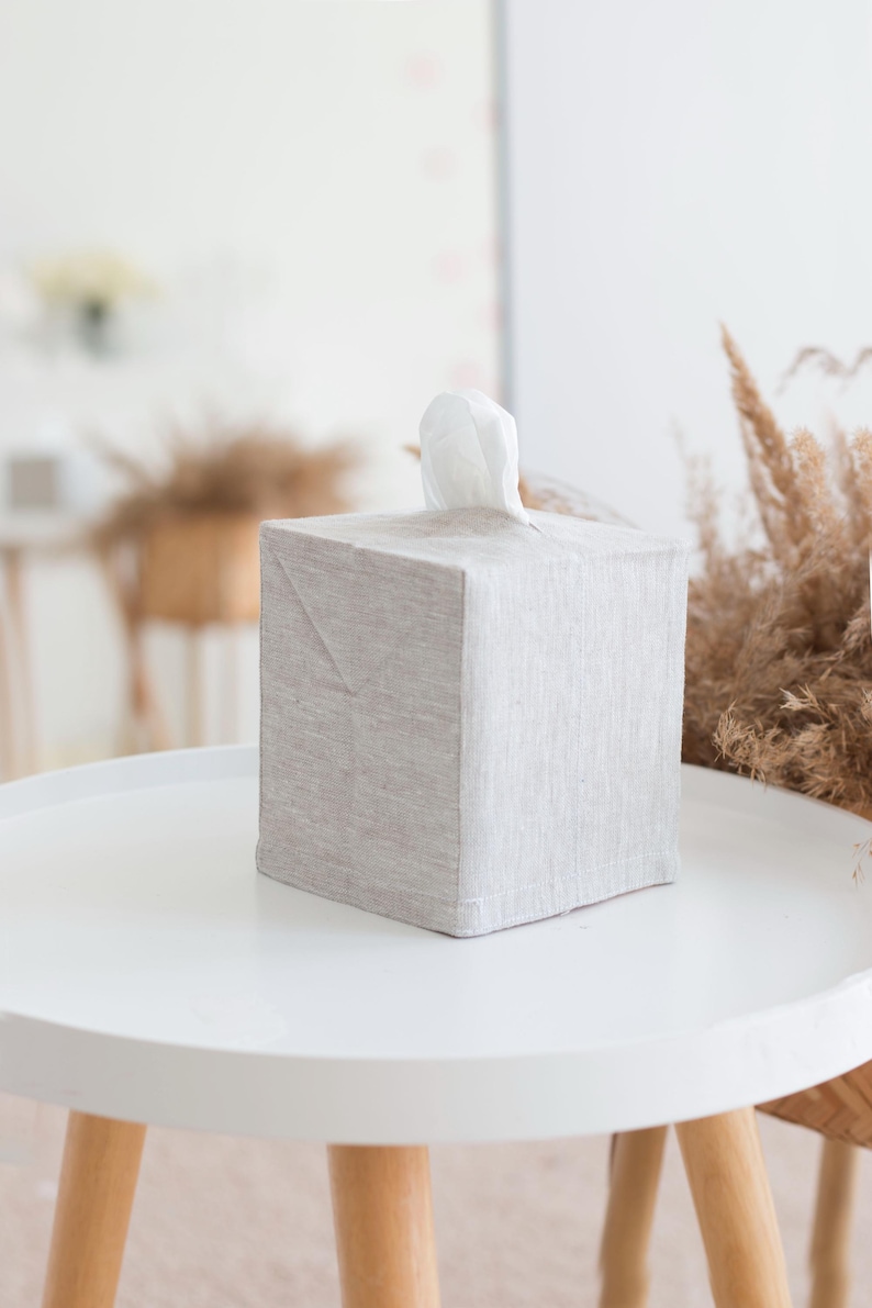 May include: A square tissue box cover made of light beige, textured fabric. The cover features a simple, clean design with a central opening at the top, through which a single white tissue is pulled upward, extending slightly above the box surface.